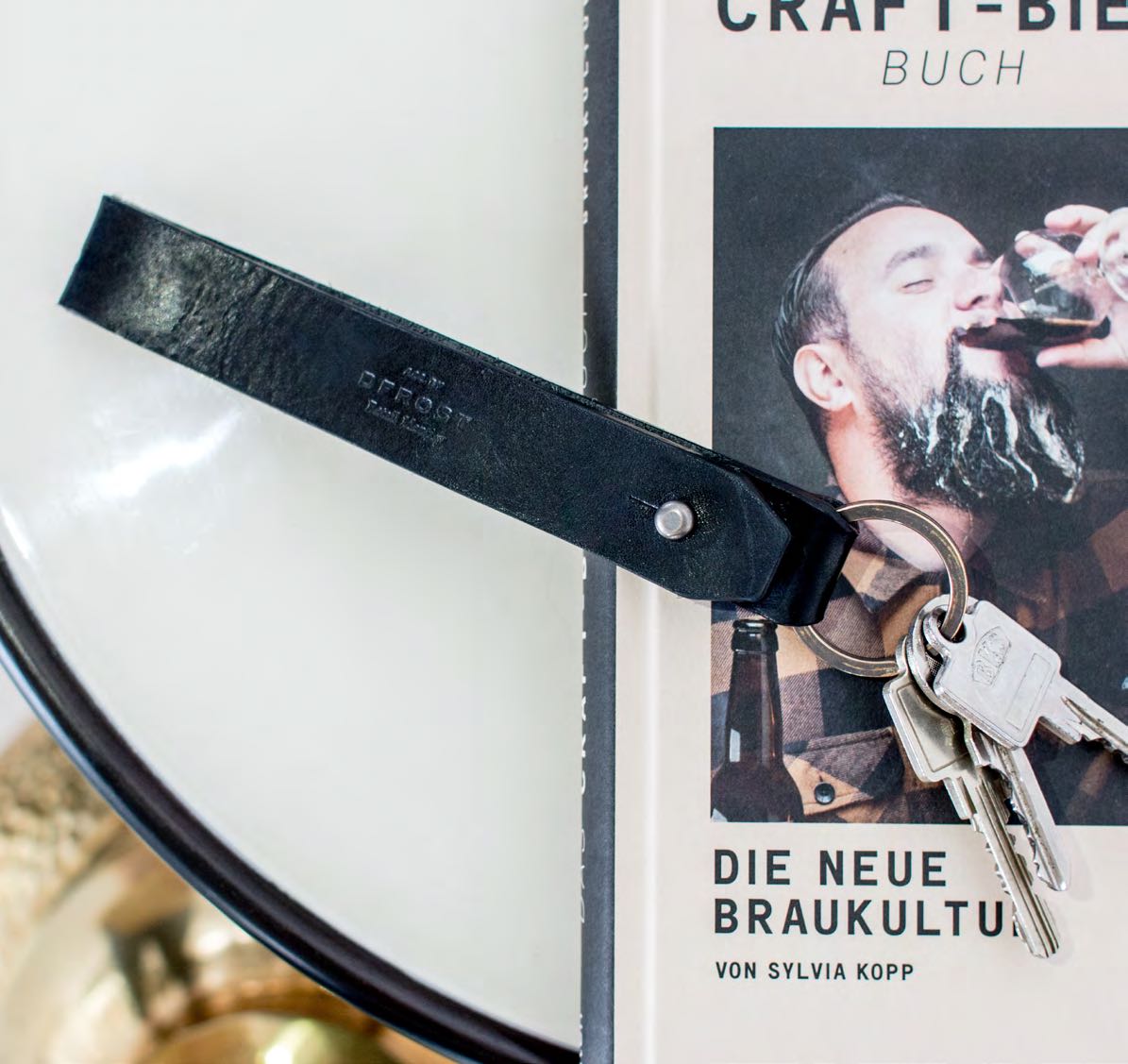 Schwarzer Schlüsselanhänger aus Leder von AYASSE, arrangiert mit einem Craft-Bier-Buch, präsentiert von MORE PIOUS STUDIOS im Rahmen des Projekts DFROST x AYASSE.